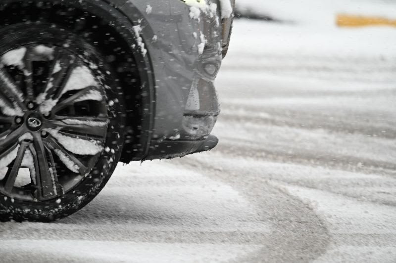 Close-up of a grey car tire and bumper covered in snow while driving on a slushy, snow-covered road during a winter storm.