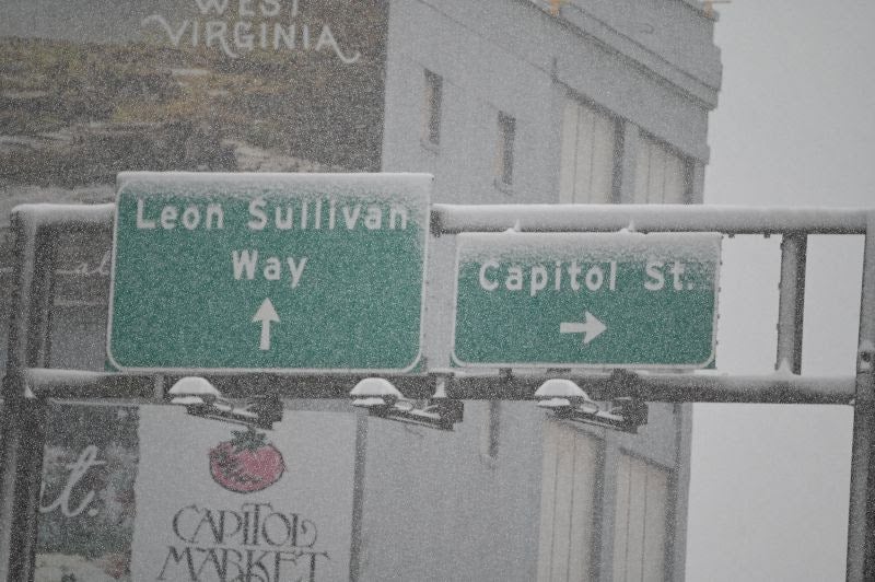 Green overhead street signs for Leon Sullivan Way and Capitol St. covered in falling snow in downtown Charleston, West Virginia