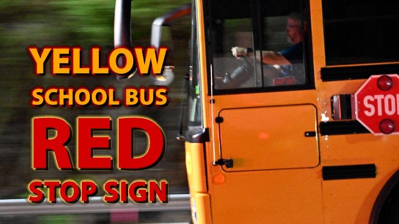 Side view of a yellow school bus driving along a road next to a guardrail and rock wall, showing the driver at the wheel and an attached red stop sign arm