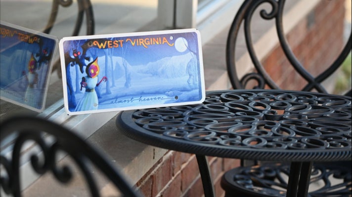 Cryptids license plate featuring mothman, bigfoot, the flatwoods monster, and other West Virginia cryptids. The plate is on an outdoor metal table and leaned against a building's window.