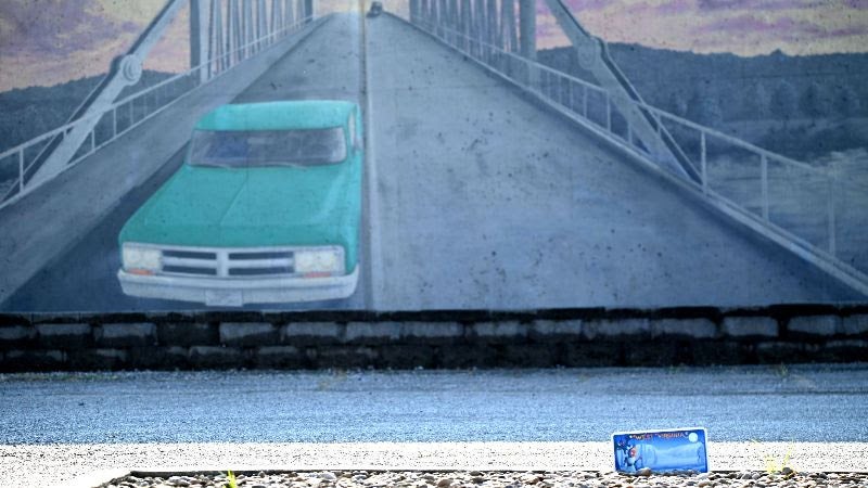 An image of the Silver Bridge Memorial Mural painted on the floodwall of Point Pleasant, West Virginia. The mural depicts a teal truck driving down the bridge. This photo also shows the new cryptids license plate in the bottom right corner.