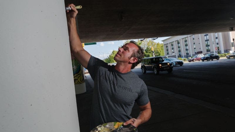 Artist Jesse Corlis painting a mural under a bridge. 
